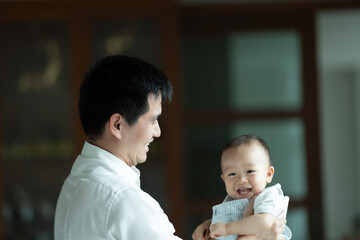 Father's Day concept, young Asian father holding a baby child with family person at home, happy childhood with love and care together, little cute son with dad and parent, parenthood lifestyle