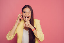 Cheerful Young Brunette Girl In Yellow Jacket Talking On Phone, Laughing After Hearing A Secret, Gesturing Silence Sign, Touching Lips By Index Finger, Girls Gossip. People Over Pastel Pink Background