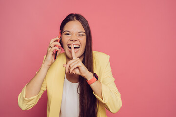 Cheerful young brunette girl in yellow jacket talking on phone, laughing after hearing a secret, gesturing silence sign, touching lips by index finger, girls gossip. People over pastel pink background