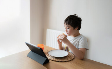 Wall Mural - Kid eating eating homemade bacon sandwiches and watching cartoon on tablet, Child boy having  breakfast and playing game online on internet with friends before go to school in the morning