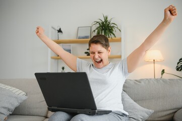 Canvas Print - Happy excited grey haired mature woman celebrating online win, using laptop, looking at screen, sitting on couch at home, middle aged female feeling amazed, surprised by unbelievable good news.