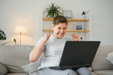 Canvas Print - Happy excited grey haired mature woman celebrating online win, using laptop, looking at screen, sitting on couch at home, middle aged female feeling amazed, surprised by unbelievable good news.