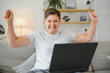 Canvas Print - Excited mature woman looking at laptop screen, reading good news in message, celebrating online lottery win, rejoicing success, overjoyed older female sitting on couch at home, using computer.