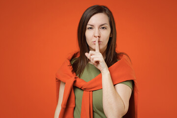 Wall Mural - Young smiling happy woman 20s wearing khaki t-shirt tied sweater on shoulders say hush be quiet with finger on lips shhh gesture isolated on plain orange background studio. People lifestyle concept.