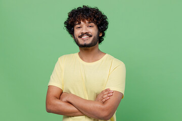 Wall Mural - Young smiling confident happy Indian man 20s in basic yellow t-shirt hold hands crossed folded look camera isolated on plain pastel light green background studio portrait. People lifestyle concept