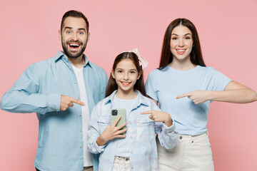 Wall Mural - Young surprised amazed parents mom dad with child kid daughter teen girl in blue clothes hold use point index finger on mobile cell phone isolated on plain pastel pink background. Family day concept.