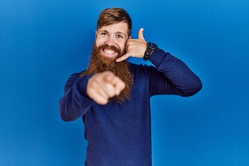 Sticker - Redhead man with long beard wearing casual blue sweater over blue background smiling doing talking on the telephone gesture and pointing to you. call me.