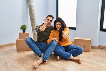 Canvas Print - Young couple sitting on the floor at new home approving doing positive gesture with hand, thumbs up smiling and happy for success. winner gesture.