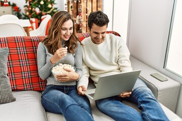 Wall Mural - Young hispanic couple watching film eating popcorn sitting on the sofa at home.