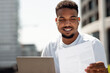 © Prostock-studio - Happy black man working with documents and laptop computer outdoors, sitting in city area and using pc, preparing report