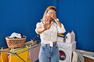 Canvas Print - Young blonde woman at laundry room covering eyes with hands and doing stop gesture with sad and fear expression. embarrassed and negative concept.