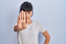 Young Hispanic Woman Wearing Casual T Shirt Over Blue Background Doing Stop Sing With Palm Of The Hand. Warning Expression With Negative And Serious Gesture On The Face.