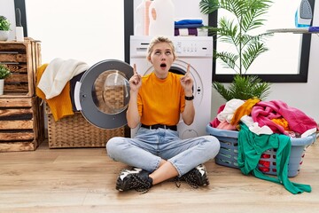 Poster - Young blonde woman doing laundry sitting by washing machine amazed and surprised looking up and pointing with fingers and raised arms.