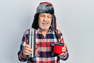 Canvas Print - Handsome mature handyman wearing winter hat with ear flaps drinking hot coffee from thermo winking looking at the camera with sexy expression, cheerful and happy face.