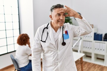 Canvas Print - Middle age doctor man at the clinic with a patient very happy and smiling looking far away with hand over head. searching concept.