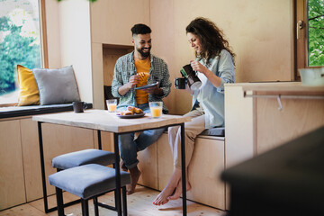 Wall Mural - Happy couple having breakfast, sitting, resting and talking indoors in a tree house, morning routine.