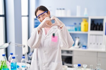 Poster - Young brunette woman working at scientist laboratory smiling in love doing heart symbol shape with hands. romantic concept.