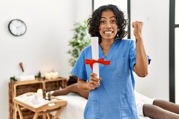 Poster - Young therapist woman holding diploma at spa wellness center screaming proud, celebrating victory and success very excited with raised arms