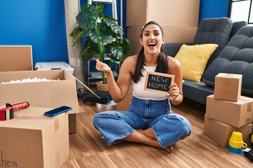 Wall Mural - Young hispanic woman holding blackboard with new home text screaming proud, celebrating victory and success very excited with raised arm