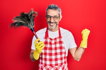 Wall Mural - Handsome middle age man with grey hair wearing apron holding cleaning duster screaming proud, celebrating victory and success very excited with raised arm