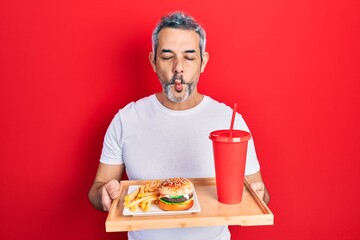 Sticker - Handsome middle age man with grey hair eating a tasty classic burger with fries and soda making fish face with mouth and squinting eyes, crazy and comical.