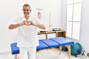 Canvas Print - Middle age hispanic therapist man working at pain recovery clinic smiling in love doing heart symbol shape with hands. romantic concept.