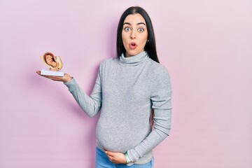 Poster - Beautiful woman with blue eyes expecting a baby, holding anatomic fetus scared and amazed with open mouth for surprise, disbelief face