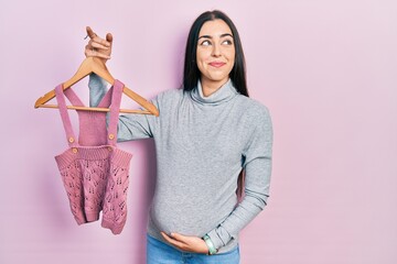 Poster - Beautiful woman with blue eyes expecting a baby holding baby clothes smiling looking to the side and staring away thinking.