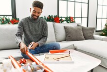 Young african american man smiling happy wrapping christmas gift sitting on the sofa at home.