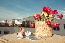 Composition Of Straw Bag With Bunch Of Pink, Yellow Flowers Tulips, Beige Sandals And Magazine On Linen Cloth On Roof.