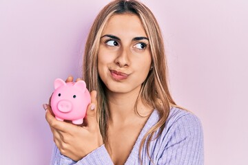 Poster - Beautiful hispanic woman holding piggy bank smiling looking to the side and staring away thinking.