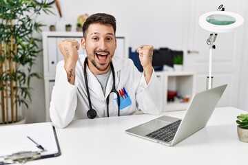 Canvas Print - Young doctor working at the clinic using computer laptop celebrating surprised and amazed for success with arms raised and open eyes. winner concept.