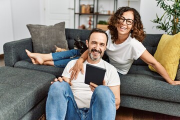 Wall Mural - Middle age hispanic couple smiling happy and using touchpad. Sitting on the sofa with dogs at home.