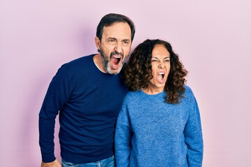 Sticker - Middle age couple of hispanic woman and man hugging and standing together angry and mad screaming frustrated and furious, shouting with anger. rage and aggressive concept.