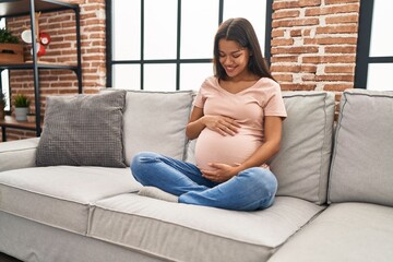 Wall Mural - Young latin woman pregnant touching belly sitting on sofa at home