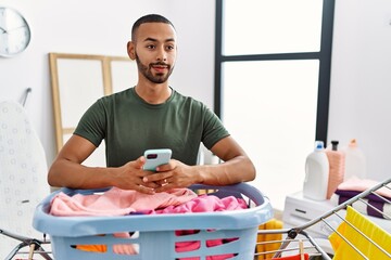 Poster - African american man doing laundry using smartphone smiling looking to the side and staring away thinking.