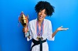 African american woman with afro hair wearing karate kimono and black belt holding trophy and medals celebrating achievement with happy smile and winner expression with raised hand