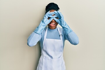 Wall Mural - African american woman with braided hair wearing cleaner apron and gloves doing ok gesture like binoculars sticking tongue out, eyes looking through fingers. crazy expression.