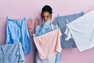 Wall Mural - African american woman with braided hair washing clothes at clothesline shocked covering mouth with hands for mistake. secret concept.