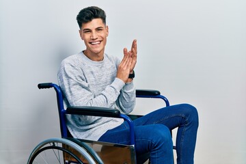 Wall Mural - Young hispanic man sitting on wheelchair clapping and applauding happy and joyful, smiling proud hands together