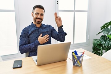 Canvas Print - Young hispanic man with beard working at the office with laptop smiling swearing with hand on chest and fingers up, making a loyalty promise oath