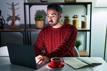 Wall Mural - Young hispanic man with beard using computer laptop at night at home puffing cheeks with funny face. mouth inflated with air, crazy expression.