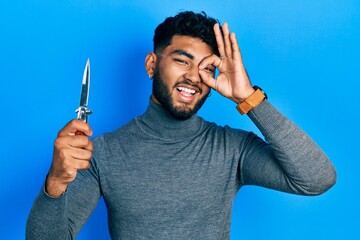 Wall Mural - Arab man with beard holding pocket knife smiling happy doing ok sign with hand on eye looking through fingers