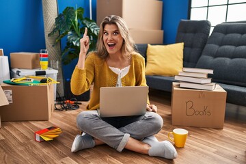 Canvas Print - Young woman sitting on the floor at new home using laptop pointing finger up with successful idea. exited and happy. number one.