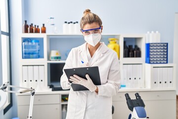 Canvas Print - Young blonde woman wearing scientist uniform and medical mask writing on clipboard at laboratory