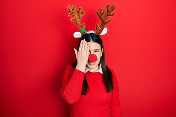 Canvas Print - Young hispanic woman wearing deer christmas hat and red nose covering one eye with hand, confident smile on face and surprise emotion.