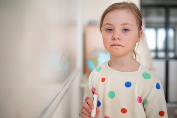 Canvas Print - Little schoolgirl with down syndrome standing in front of whiteboard and looking at camera.