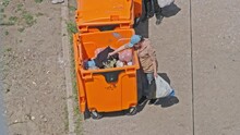 A Homeless Woman Digs In A Trash Can On The Street. View Of A Tramp Rummaging In A Rubbish Bin. Needy Homeless Woman Looking For Food In The Garbage. Ukraine