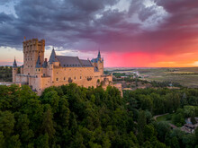 Sunset Over The Alcázar Of Segovia Medieval Castle In Castile And León, Spain. Rising Out On A Rocky Crag Above The Confluence Of Two Rivers With Majestic Red, Orange, Yellow As The Sun Paints The Sky