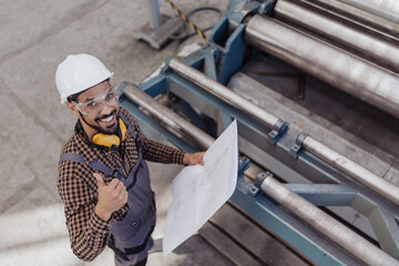 Wall Mural - High angle view of heavy industry worker with safety headphones and hard hat in industrial factory holding blueprints